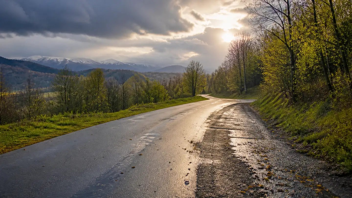Pusta mokra droga wśród zielonych wzgórz z łatanym poboczem i śnieżnymi górami w oddali - Kormoran All Season SUV na zmienną drogę przez rok